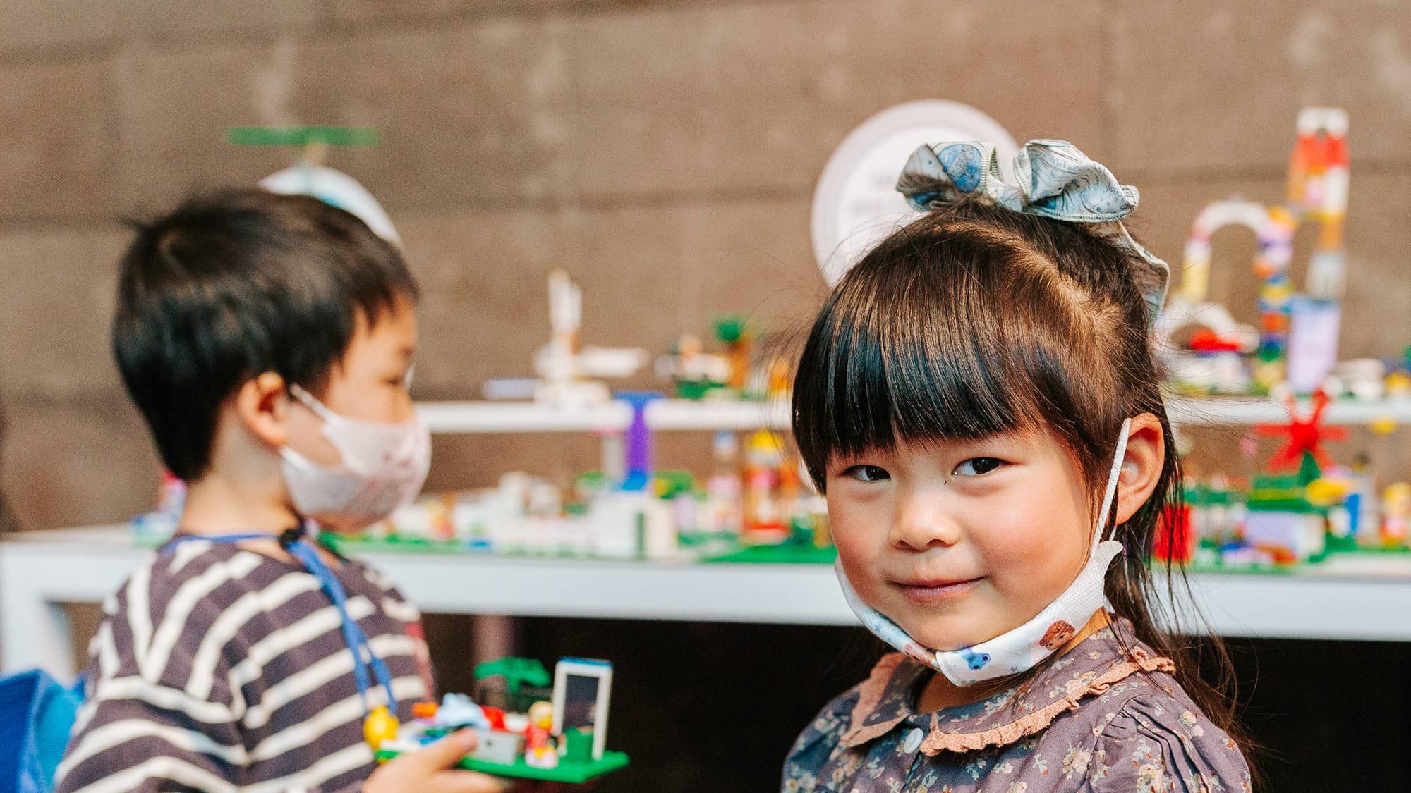 A young girl with straight black hair looking at the camera, playing with bright coloured LEGO blocks with a young boy in the background