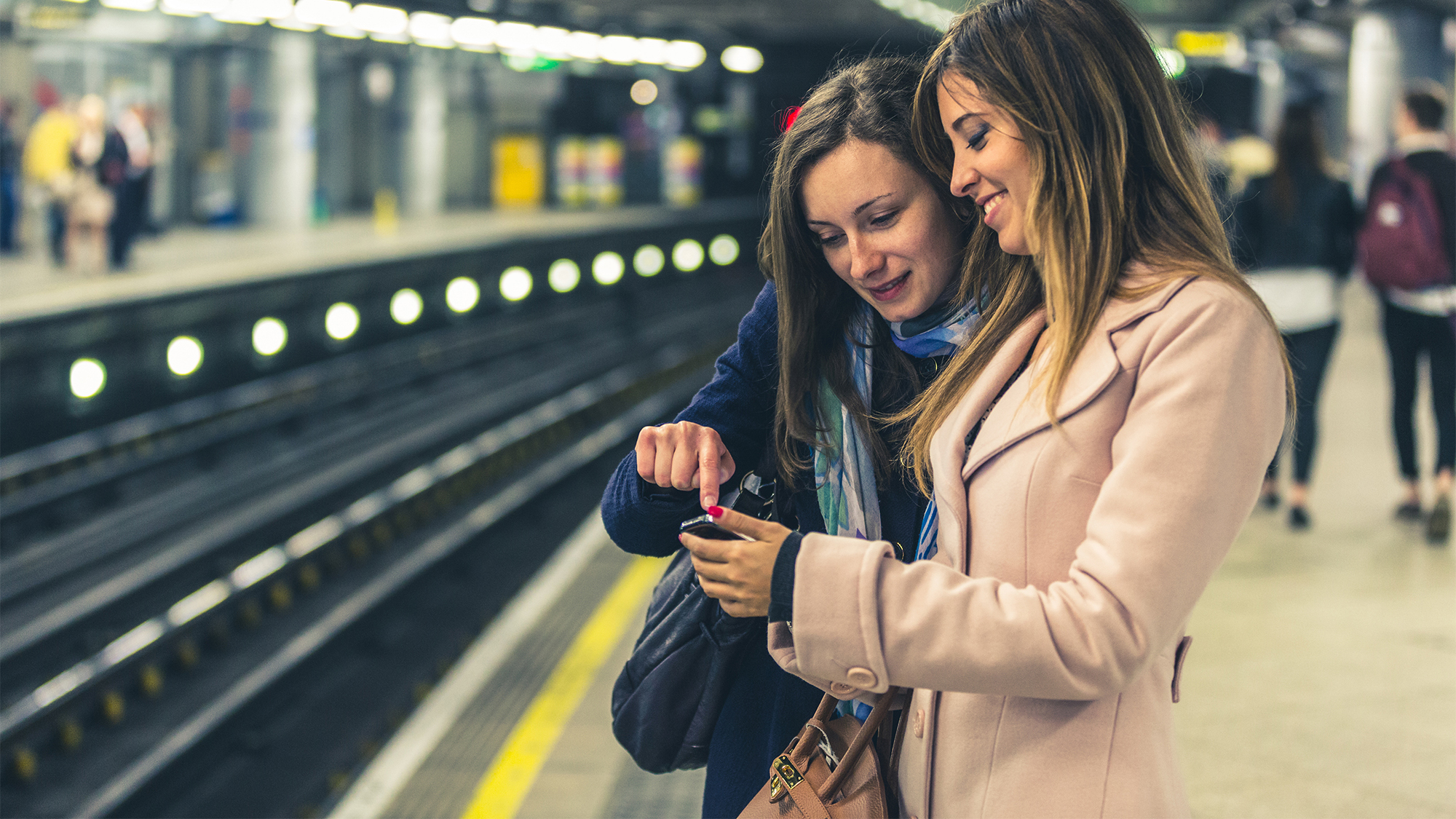 Two women use their phone while waiting on a train platform.
