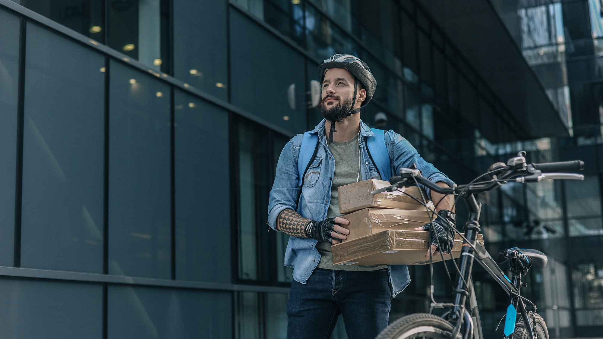 Man in street with bicycle wearing helmut and gloves holding boxes and looking ahead to deliver them