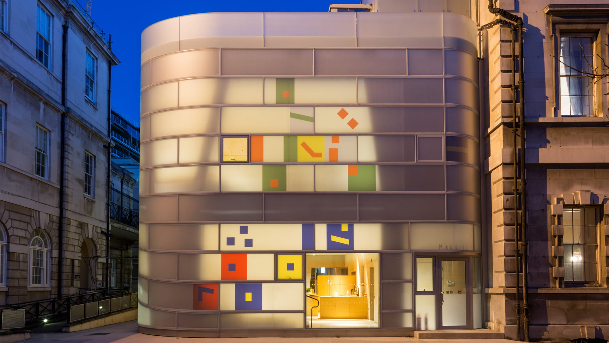 The curved, opaque glass building illuminated at night with sporadic coloured fragments on the fa?ade.