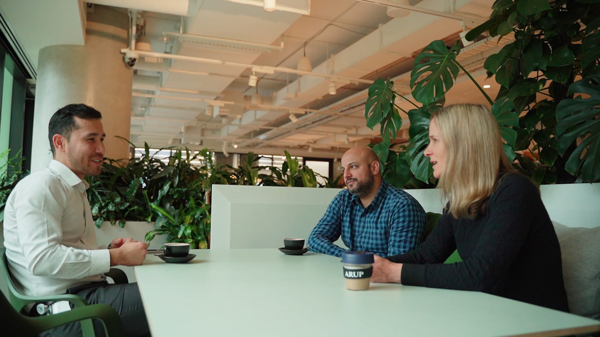 Two men and a woman sitting at a table talking, surrounded by plants and timber, in a modern work office. 