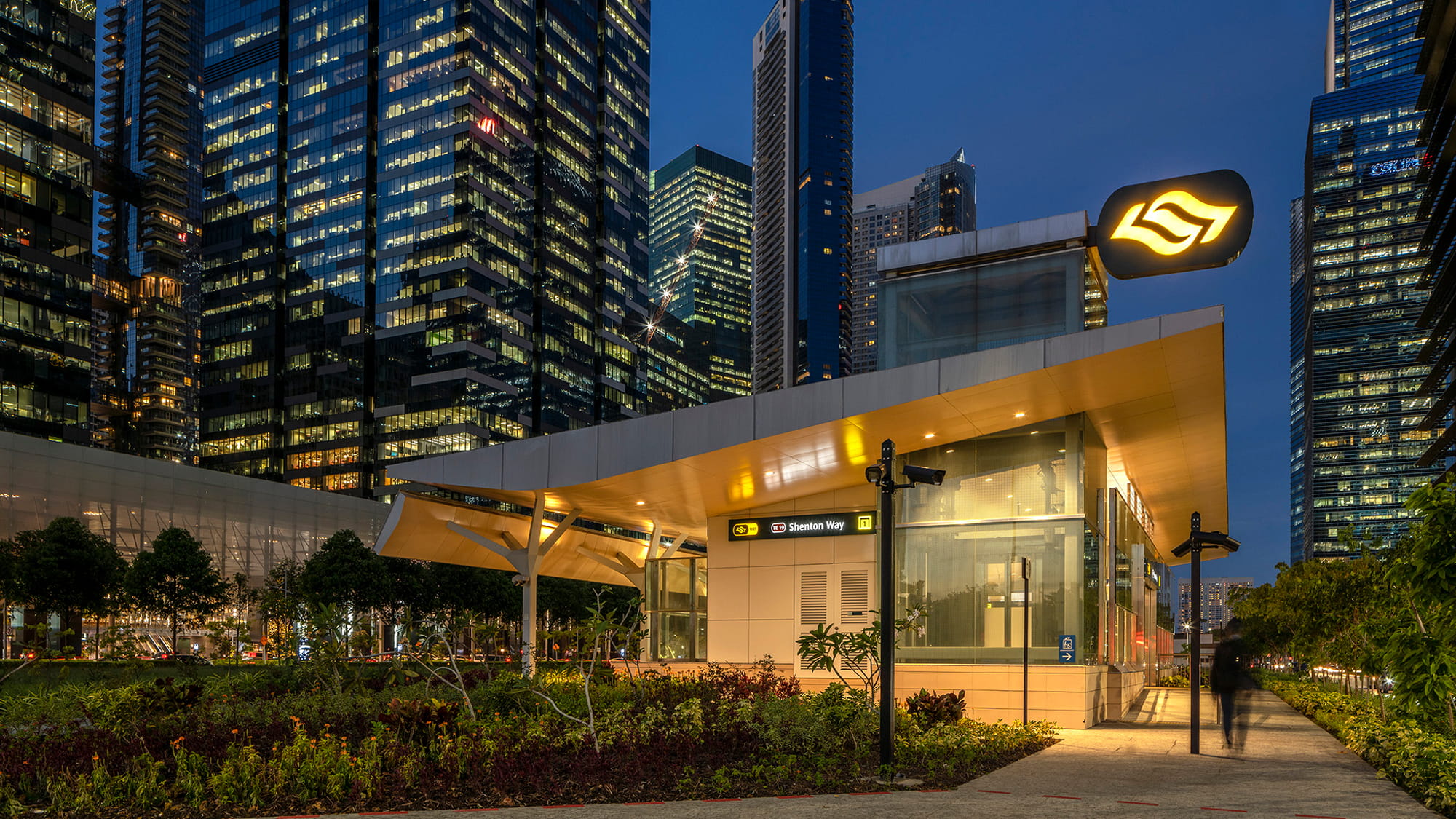 Night time view of train station building at ground level with city skyscrapers surrounding it. Green spaces, footpaths.